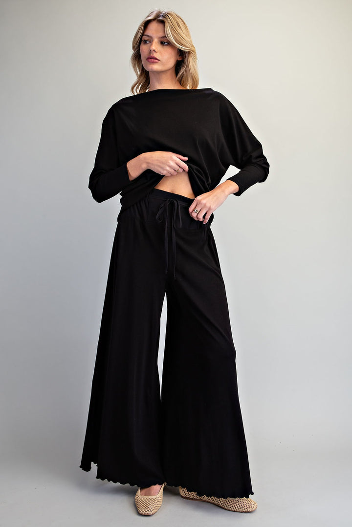 A woman modeling a black two-piece loungewear set consisting of a long-sleeve cropped top and matching wide-leg drawstring pants, paired with woven espadrille sandals against a light gray studio background.