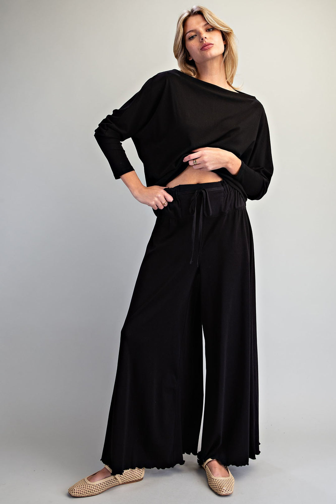 A woman modeling a black two-piece outfit consisting of a long-sleeve cropped top and matching wide-leg drawstring pants, paired with beige woven flat shoes against a light gray studio background.