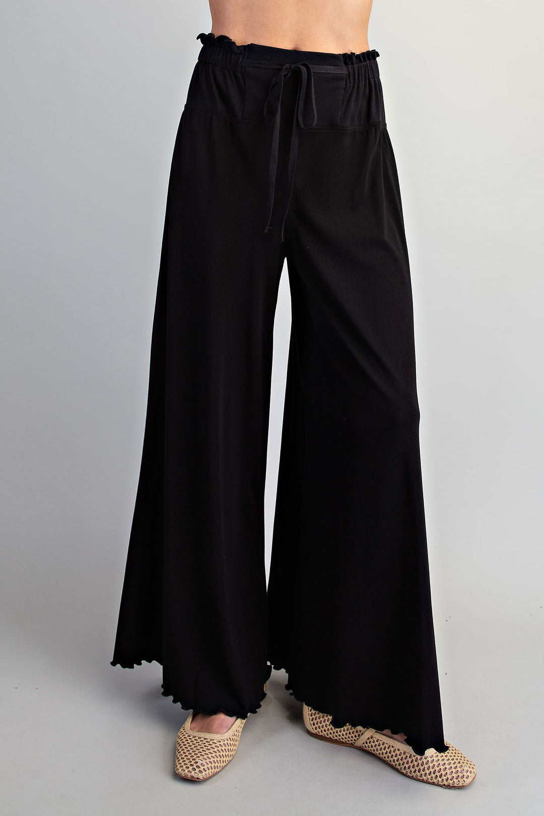 Black wide-leg pants with an elastic drawstring waist and scalloped hems, shown on a model wearing beige woven flat shoes against a light gray studio background.