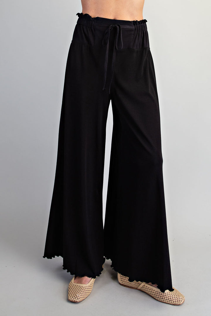 Black wide-leg pants with an elastic drawstring waist and scalloped hems, shown on a model wearing beige woven flat shoes against a light gray studio background.