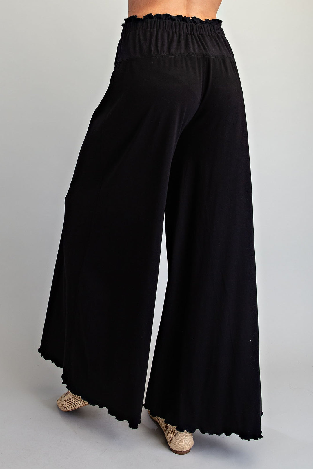 Black wide-leg pants with an elastic waistband and scalloped hem, shown on a model from the back against a light gray studio background.