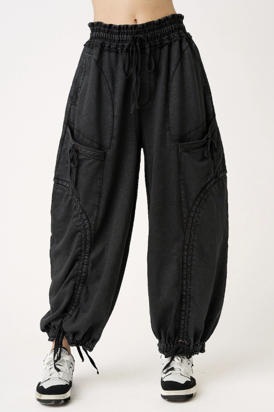 Black baggy harem-style pants with an elastic drawstring waist, large side cargo pockets, and elastic ankle cuffs, shown on a model from the waist down against a light gray studio background.