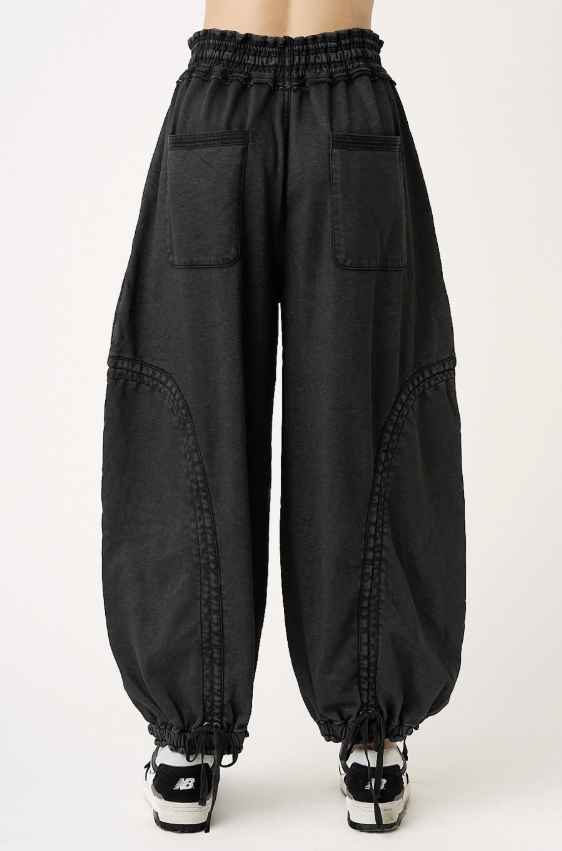 Black baggy harem-style pants with an elastic waistband, large front patch pockets, and drawstring ankle cuffs, shown on a model from the back against a light gray studio background.