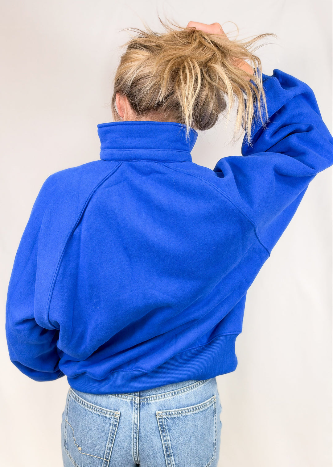 Woman shown from back wearing cobalt blue sweatshirt with jeans on plain background