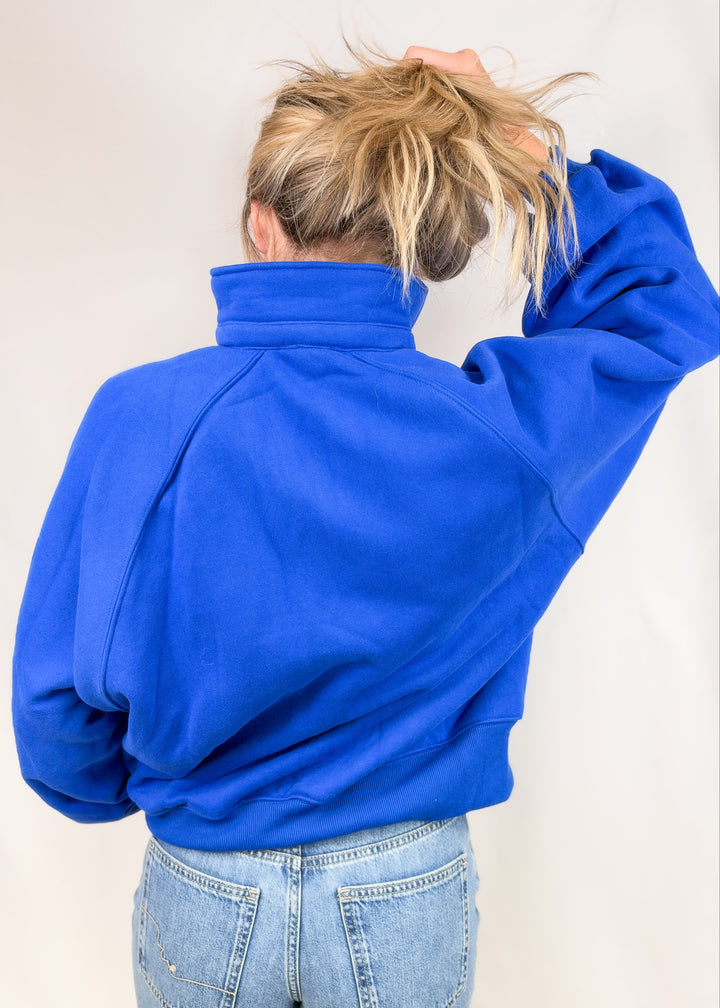 Woman shown from back wearing cobalt blue sweatshirt with jeans on plain background