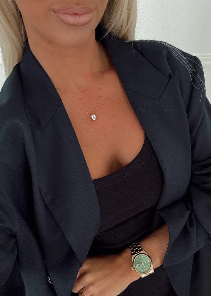A close-up lifestyle shot of a woman wearing a black satin blazer over a black top, accessorized with a delicate silver necklace with a small square gemstone and a gold watch with a green dial.