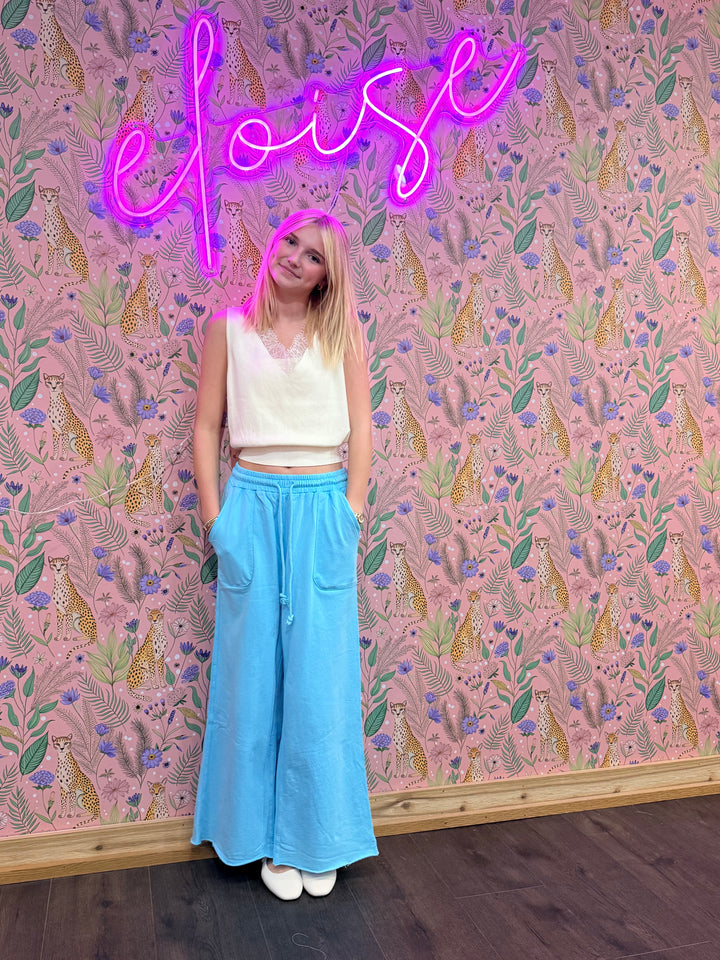Person wearing a white top and blue pants standing in front of a patterned wall with a neon sign.