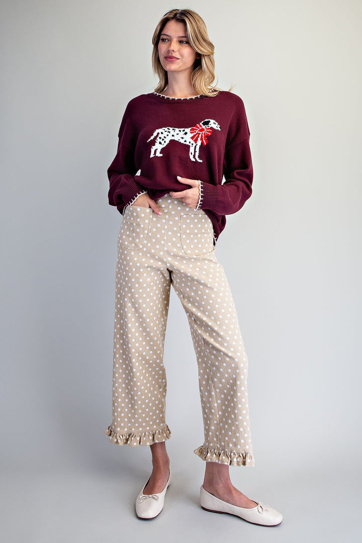 A woman modeling a burgundy long-sleeve sweater with a white and red Dalmatian dog graphic and a red bow, paired with beige wide-leg pants featuring white polka dots and ruffled hems, and white ballet flats, against a light gray studio background.