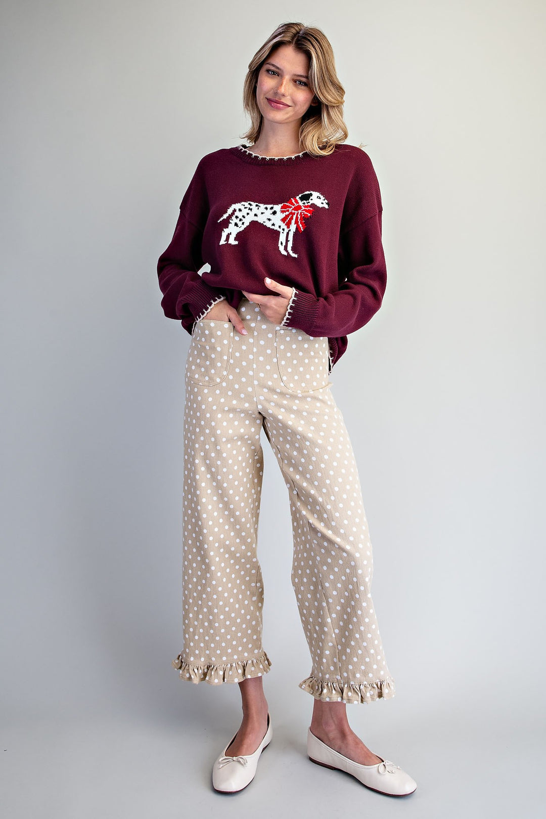 A woman modeling a burgundy long-sleeve sweater with a white dalmatian and red bow graphic on the chest, paired with beige wide-leg pants featuring white polka dots and ruffled hems, against a light gray studio background.