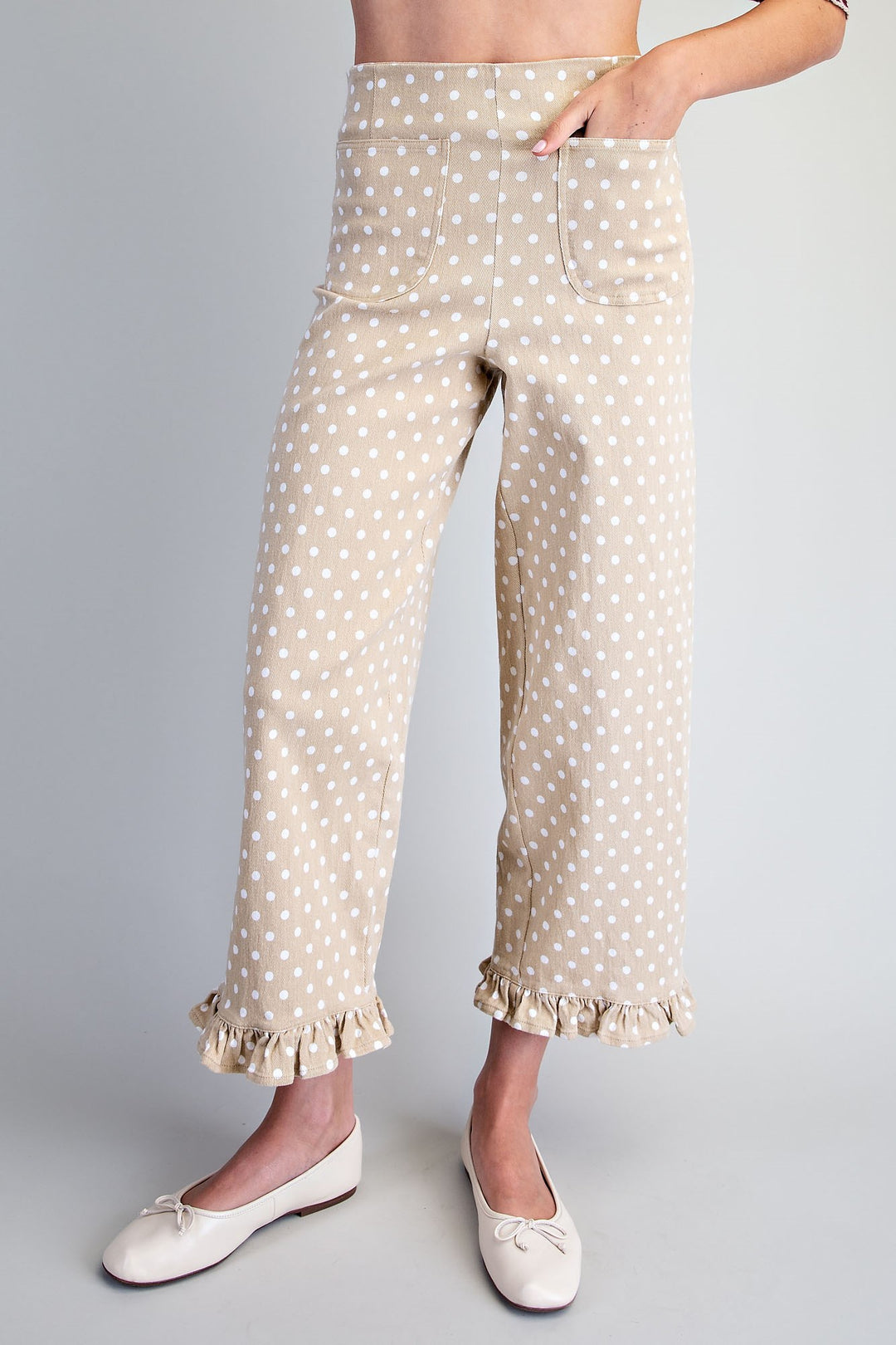 A person wearing beige cropped pants with a white polka dot pattern and ruffled hems, paired with white ballet flats, shown against a light gray studio background.