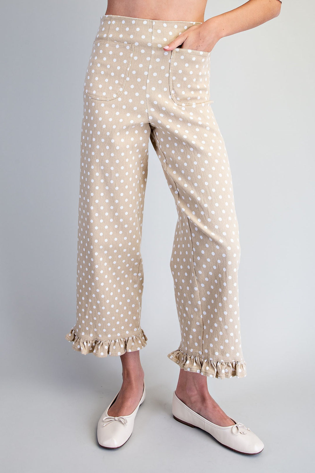 A person wearing beige cropped pants with a white polka dot pattern and ruffled hems, paired with white ballet flats, shown against a light gray studio background.