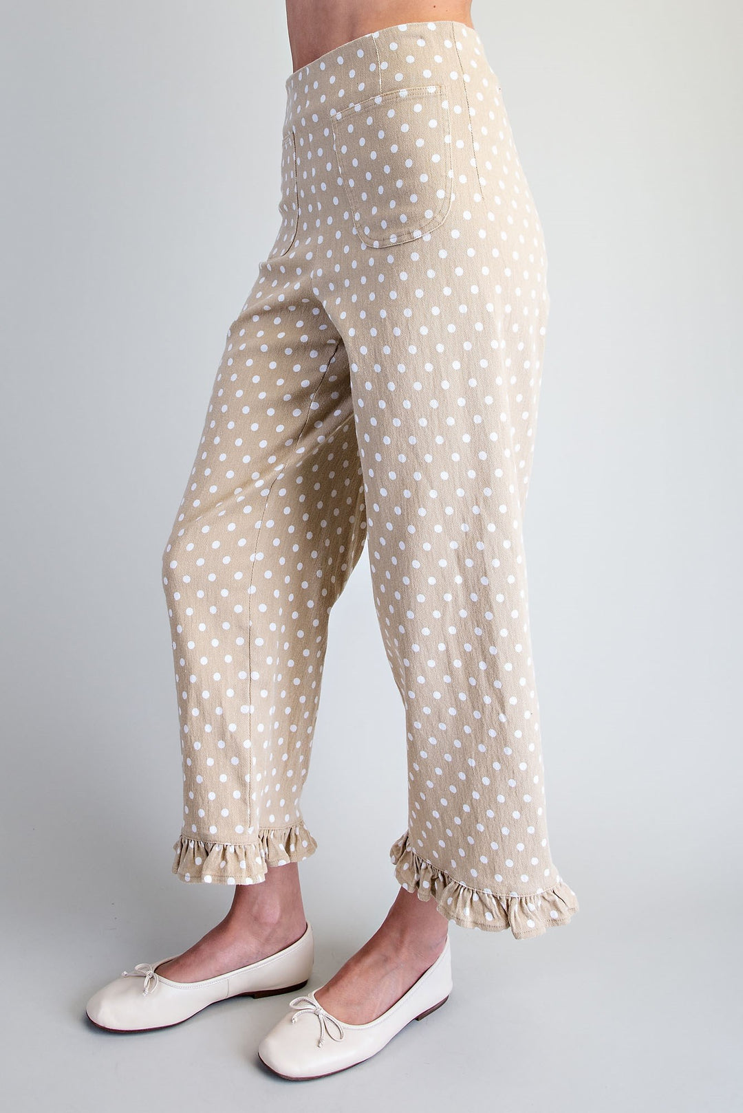 A person wearing beige pants with a white polka dot pattern and ruffled hems, paired with white ballet flats, shown from the waist down against a light gray studio background.