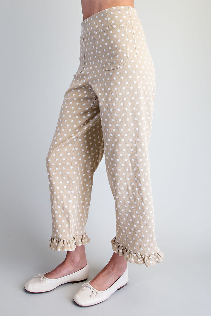 A person wearing beige pants with a white polka dot pattern and ruffled hems, paired with white ballet flats, shown from the waist down against a light gray studio background.