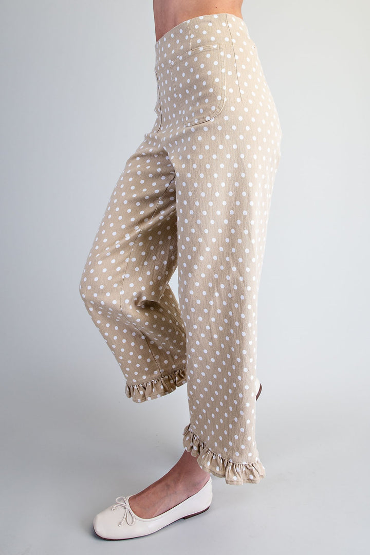 A person wearing beige wide-leg pants with a white polka dot pattern and ruffled hems, paired with white ballet flats, shown from the waist down against a light gray studio background.