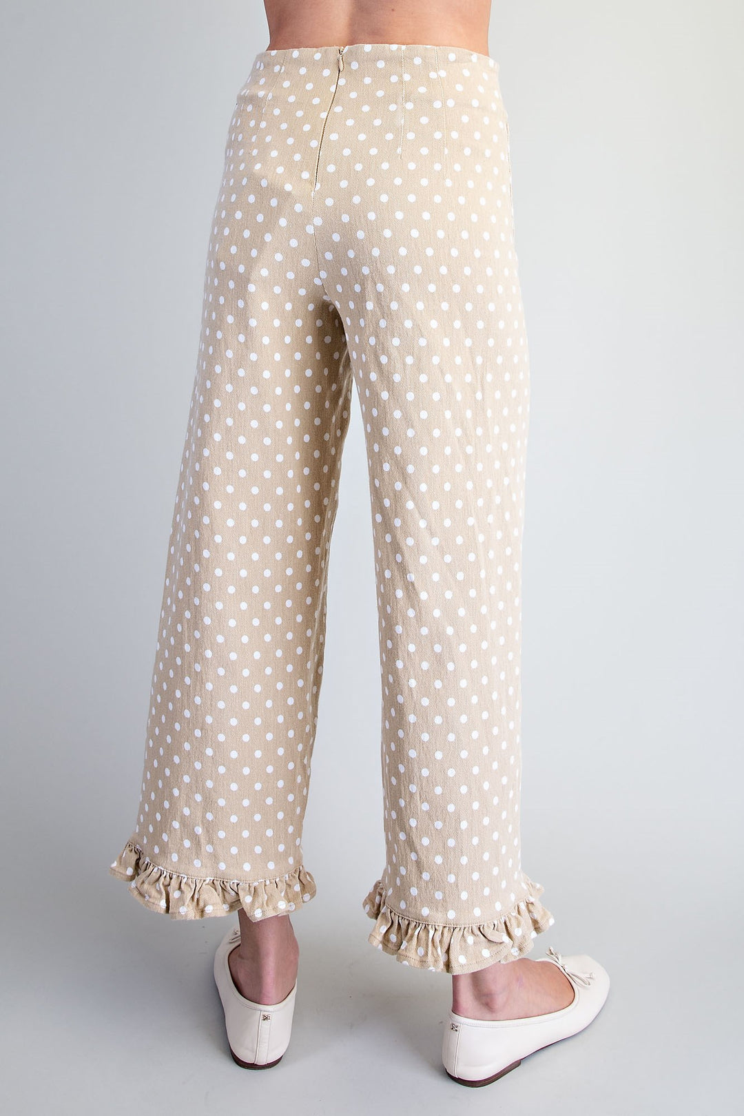 A person shown from the back wearing beige wide-leg pants with white polka dots and ruffled hems, paired with white flats, against a light gray studio background.
