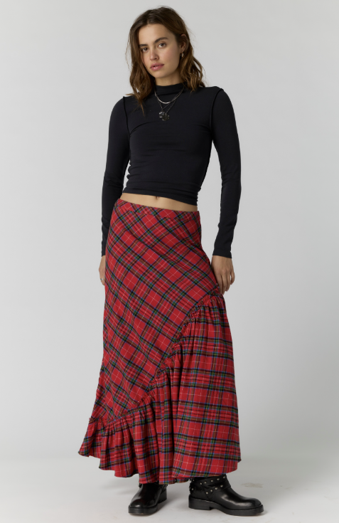 A model wearing a red and black plaid wrap maxi skirt with a ruffled hem, paired with a black long-sleeve crop top and black boots, against a light gray studio background.
