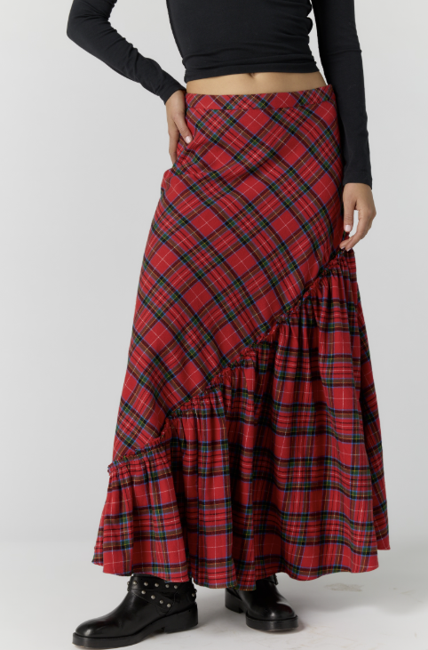 A model wearing a red and black plaid maxi skirt with a diagonal wrap design and ruffled hem, styled with a black long-sleeve crop top and black boots.