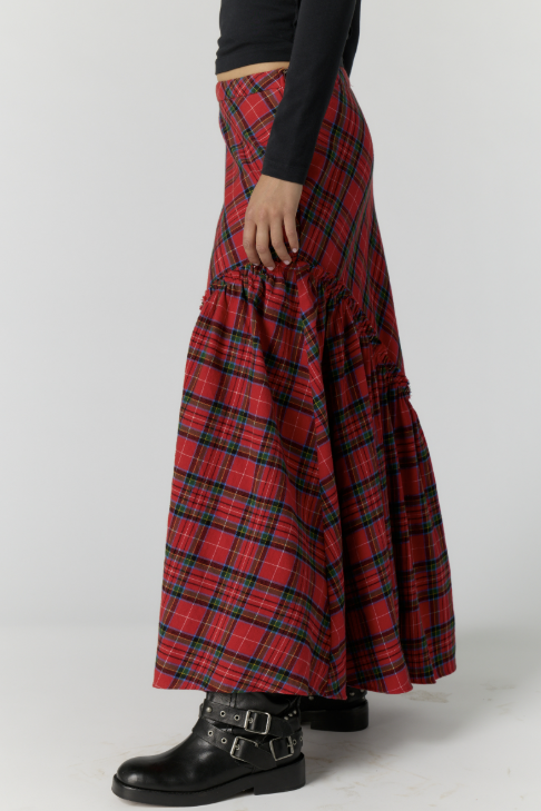 A person wearing a long red tartan plaid skirt with black and green stripes, paired with a black long-sleeve top and black buckle boots, shown from the waist down against a light gray studio background.