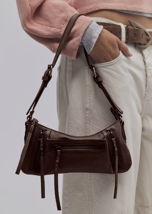 Woman holding brown leather crossbody purse wearing salmon color sweatshirt and beige pants on plain background