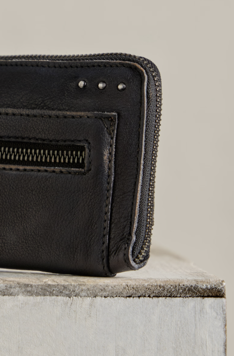 Close-up of a black leather zip-around wallet with a front slip pocket and metal studs, resting on a light gray stone block against a neutral beige background.