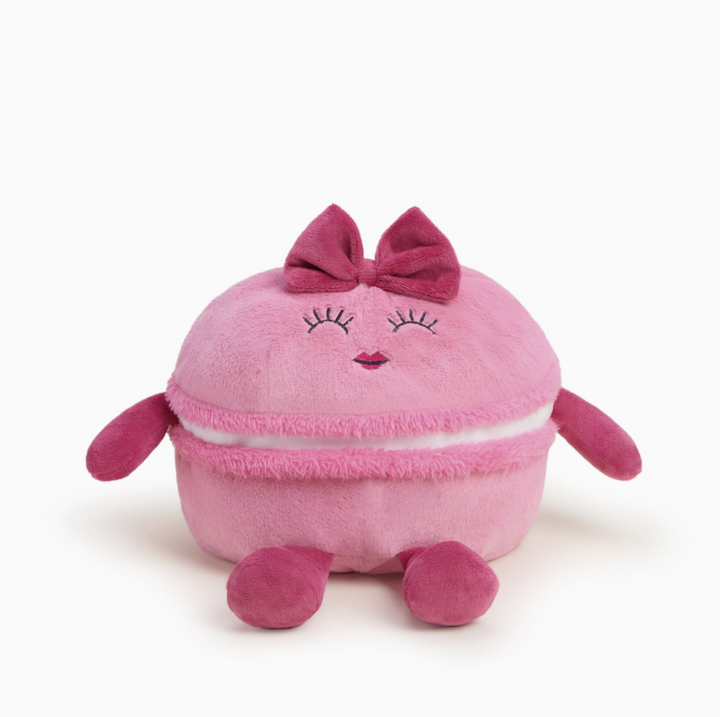 A pink plush macaron toy with embroidered closed eyes, a pink bow on top, and small pink arms and legs, photographed on a clean white background.