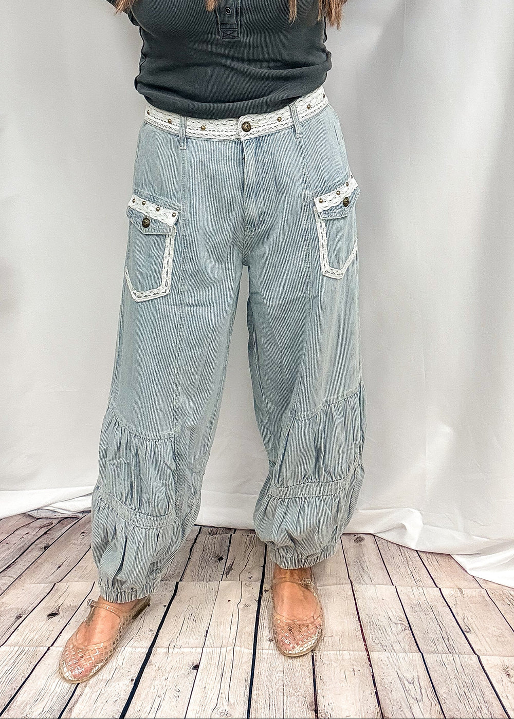 A woman stands in front of a white fabric backdrop wearing light-wash wide-leg denim pants with gathered ruffle details at the hems and pockets, paired with a dark green long-sleeve henley top and beige flat shoes.