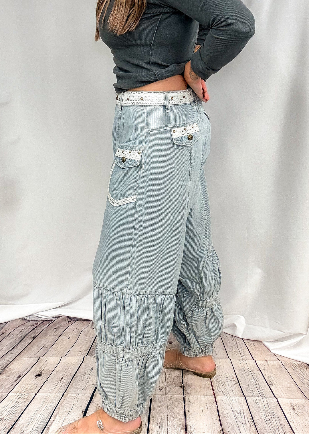 A woman modeling a pair of light-wash, high-waisted, wide-leg denim pants with decorative white embroidery and ruffle details, paired with a dark gray long-sleeve crop top, standing in front of a white fabric backdrop.