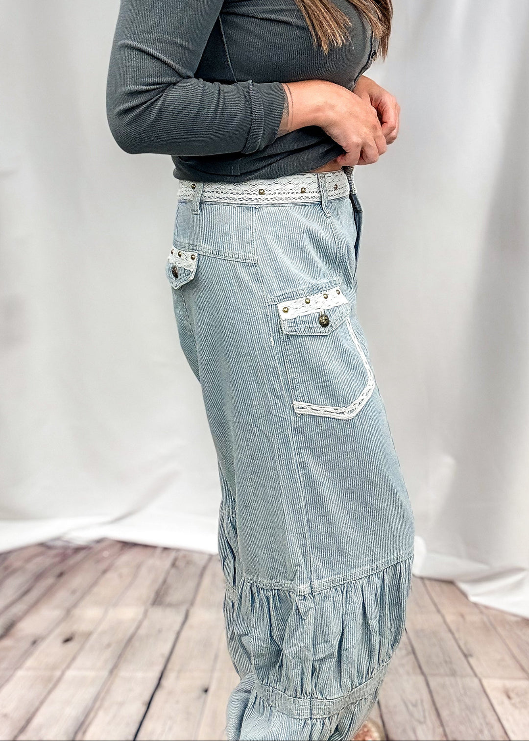A woman modeling a light-wash denim maxi skirt with ruffle details and silver rhinestone trim, paired with a dark gray long-sleeve top, standing in front of a white fabric backdrop.