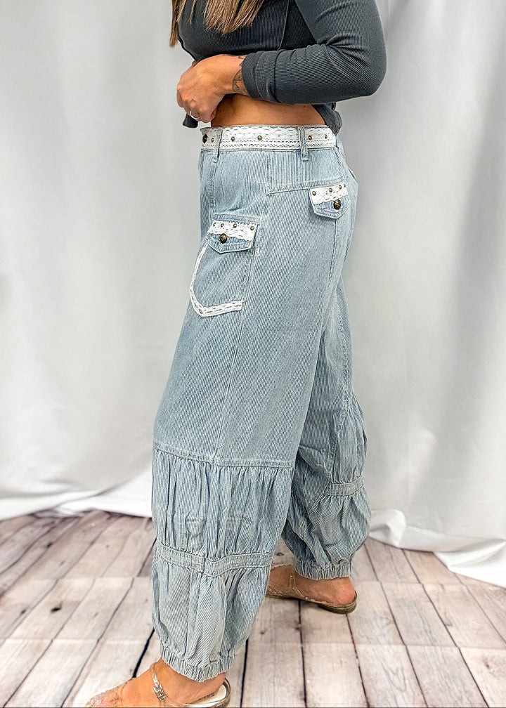 A woman modeling a pair of light-wash wide-leg denim jeans with ruffled details and a white belt, paired with a dark gray long-sleeve crop top, standing in front of a white fabric backdrop.