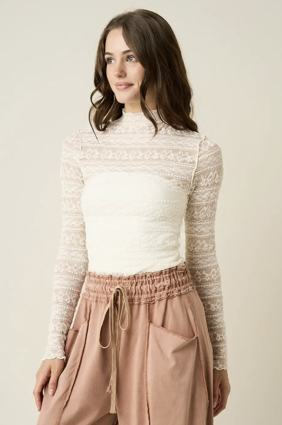A woman wearing a cream lace long-sleeve mock-neck top and dusty rose drawstring pants, photographed against a plain light beige studio background.