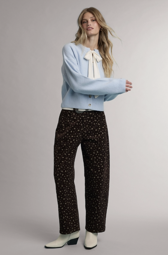 A model stands against a light gray studio background wearing a light blue cropped cardigan with a white bow at the neckline, dark brown patterned trousers, and white heeled boots.