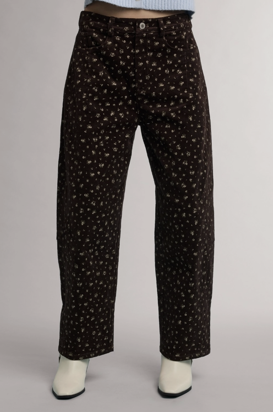 A person wearing dark brown wide-leg pants with a small white floral pattern, paired with white boots and a light blue top, shown against a neutral gray background.