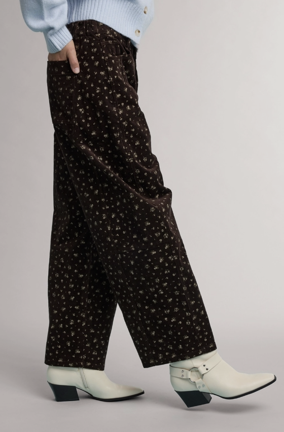 A person wearing dark brown wide-leg trousers with a small white floral pattern, styled with a light blue cardigan and white ankle boots against a neutral gray studio background.