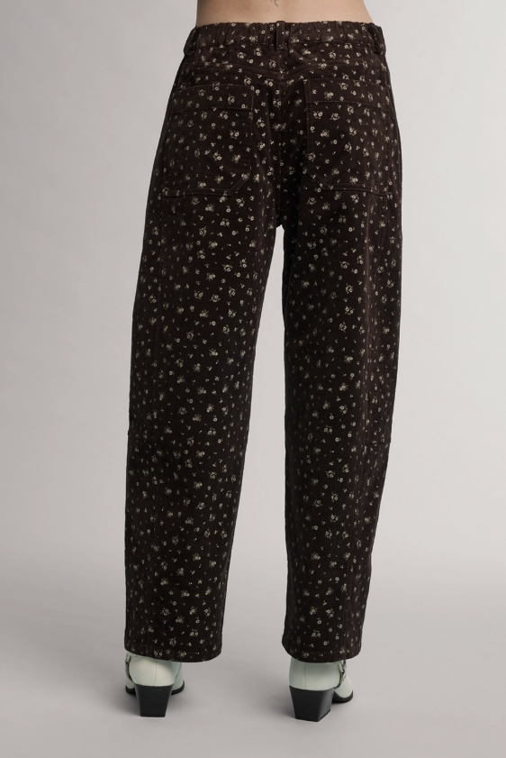 A pair of wide-leg trousers with a small white floral pattern on a dark brown base, shown from the back on a model wearing black and white heels against a light gray background.