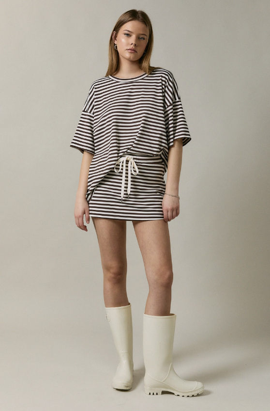 A woman modeling an oversized short-sleeve mini dress with horizontal black and off-white stripes, featuring a white drawstring tie at the waist. She is wearing off-white rain boots against a plain, light beige studio background.