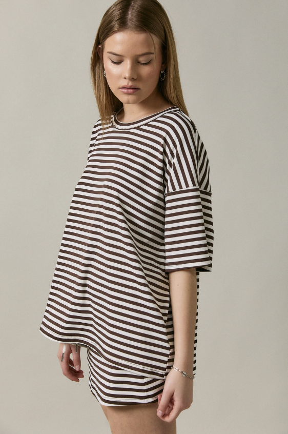 A woman wearing an oversized brown and white horizontally striped t-shirt dress, photographed against a solid light gray studio background.