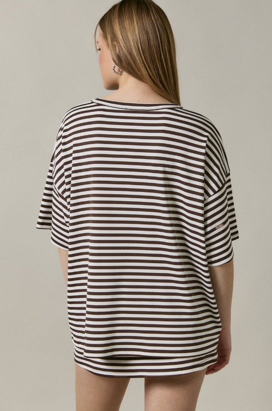 A woman shown from the back wearing an oversized short-sleeve T-shirt with horizontal dark brown and off-white stripes, set against a plain light gray studio background.
