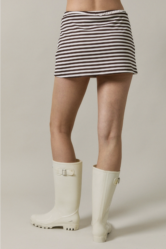 A model from the waist down wearing a brown and white striped mini skirt and off-white rubber rain boots, photographed against a neutral light gray studio background.