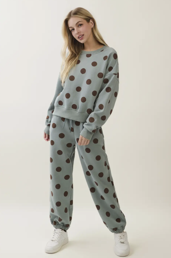 A woman modeling a matching two-piece lounge set consisting of a sweatshirt and jogger pants in a dusty sage green color with a dark brown polka dot pattern, paired with white sneakers against a plain off-white studio background.