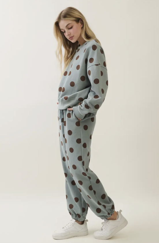 A woman modeling a matching lounge set consisting of a sweatshirt and jogger pants in a dusty sage green color with a dark brown polka dot pattern, paired with white sneakers, against a plain off-white studio background.