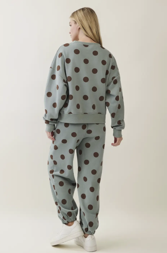 A woman shown from the back wearing a matching sage green sweatshirt and jogger pants set with a dark brown polka dot pattern, paired with white sneakers, against a plain light beige studio background.