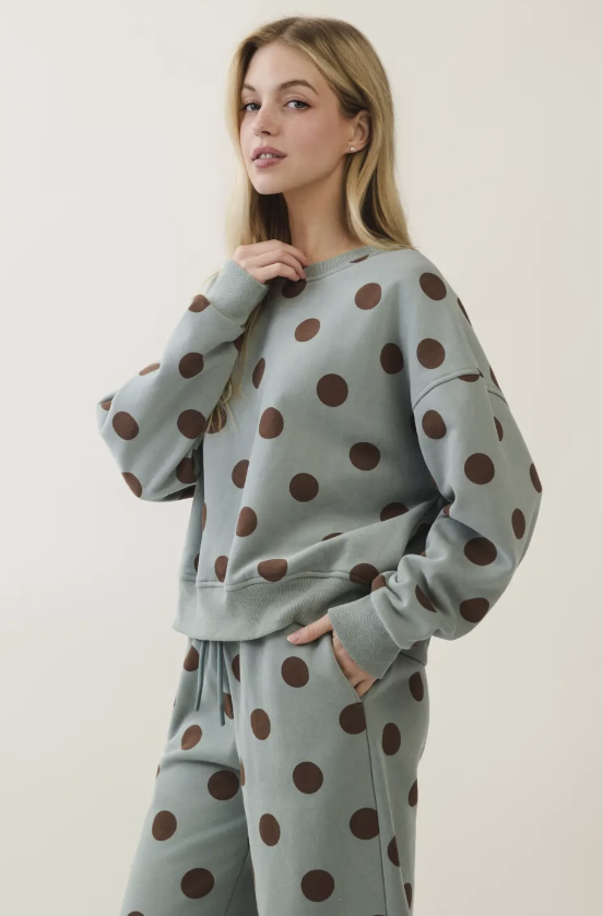 A woman modeling a matching lounge set consisting of a sweatshirt and sweatpants in a dusty sage green color with a large dark brown polka dot pattern, against a plain off-white background.