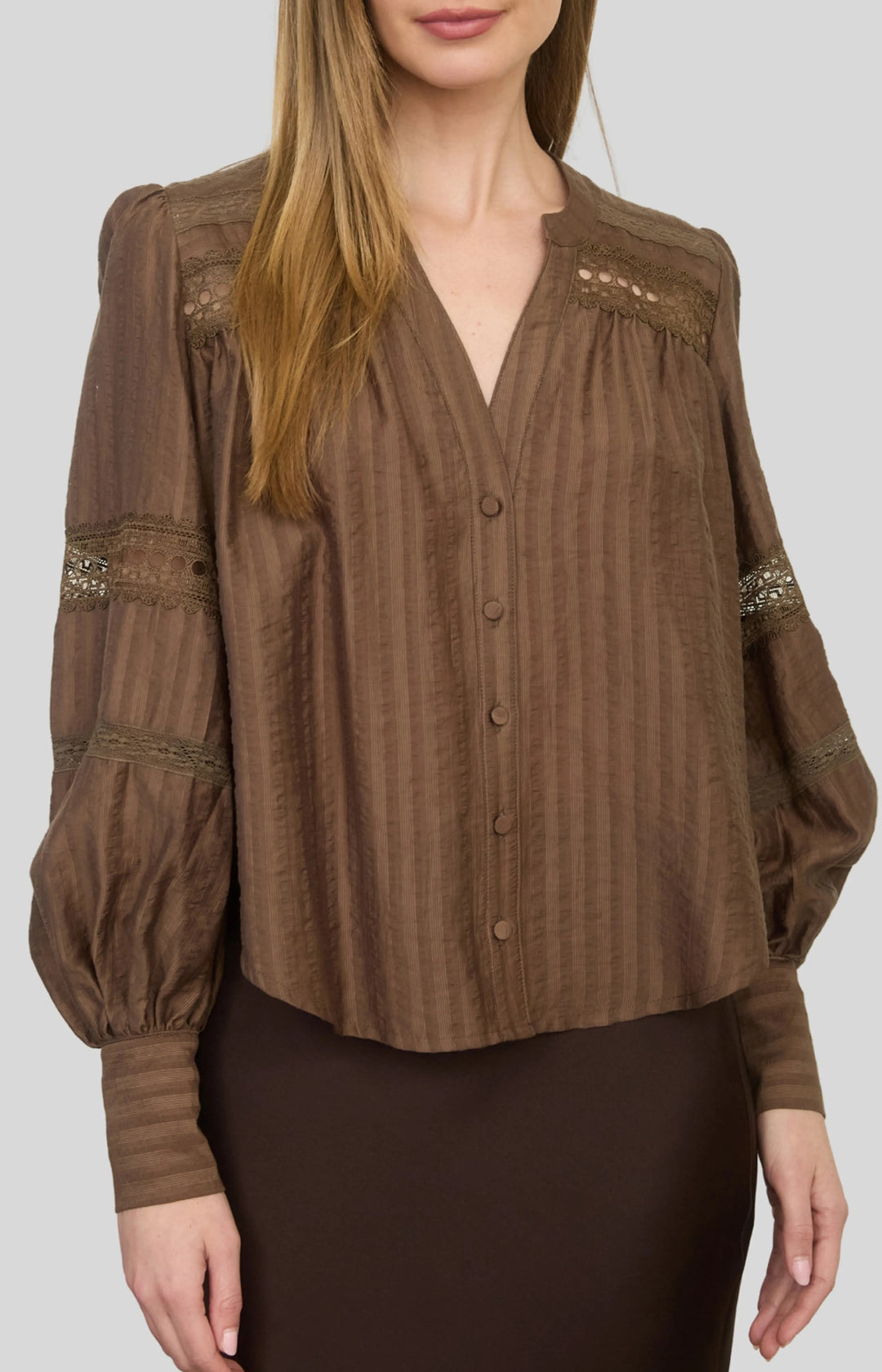 A woman wearing a brown long-sleeve button-up blouse with vertical stripes, lace-like shoulder details, and lace cuffs, paired with a dark brown skirt, against a solid gray background.