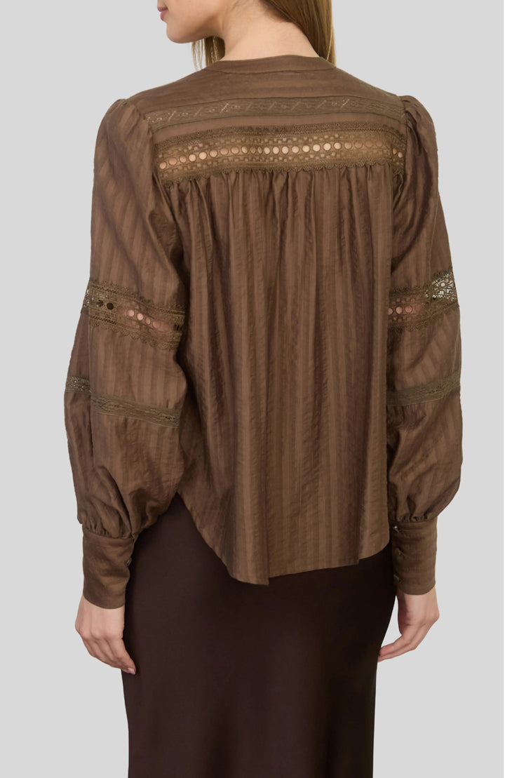 A woman wearing a brown long-sleeve blouse with lace and embroidery details on the back, paired with a dark brown skirt, shown against a solid gray background.