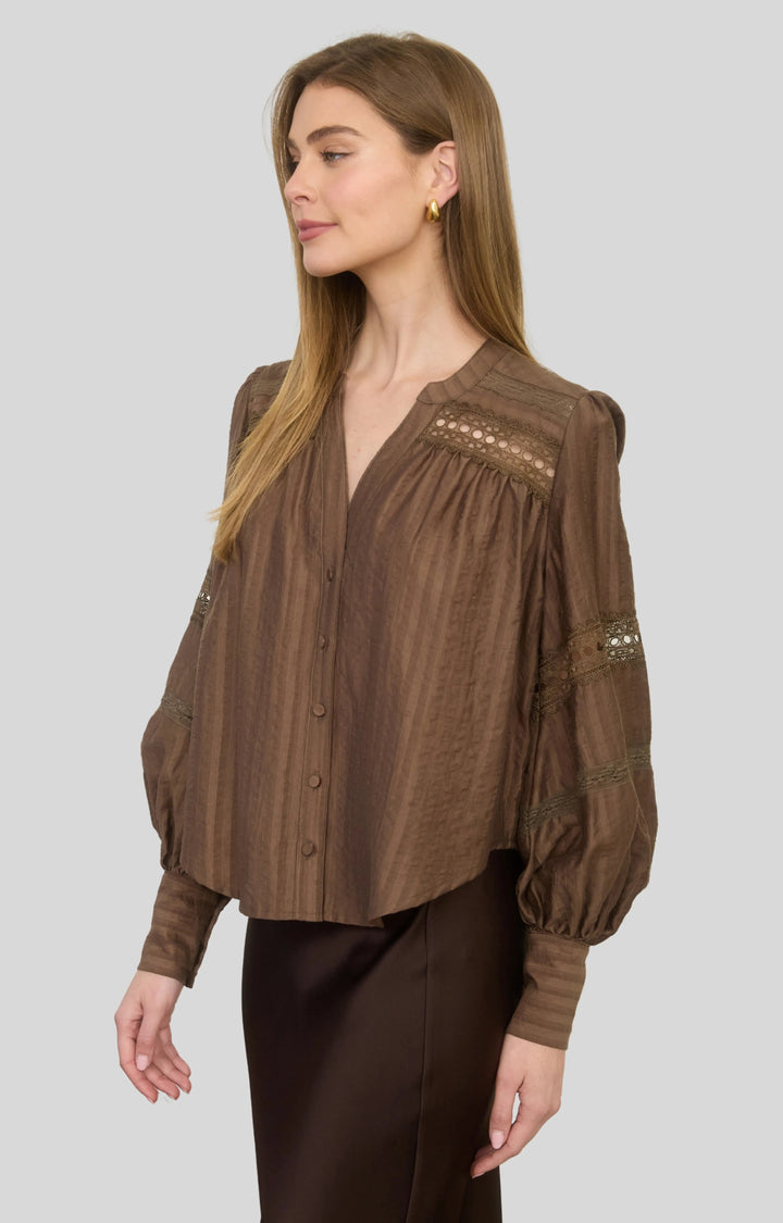 A woman modeling a brown long-sleeve blouse with vertical texture, button front, and lace detailing on the chest and sleeves, paired with dark brown pants, against a solid gray background.