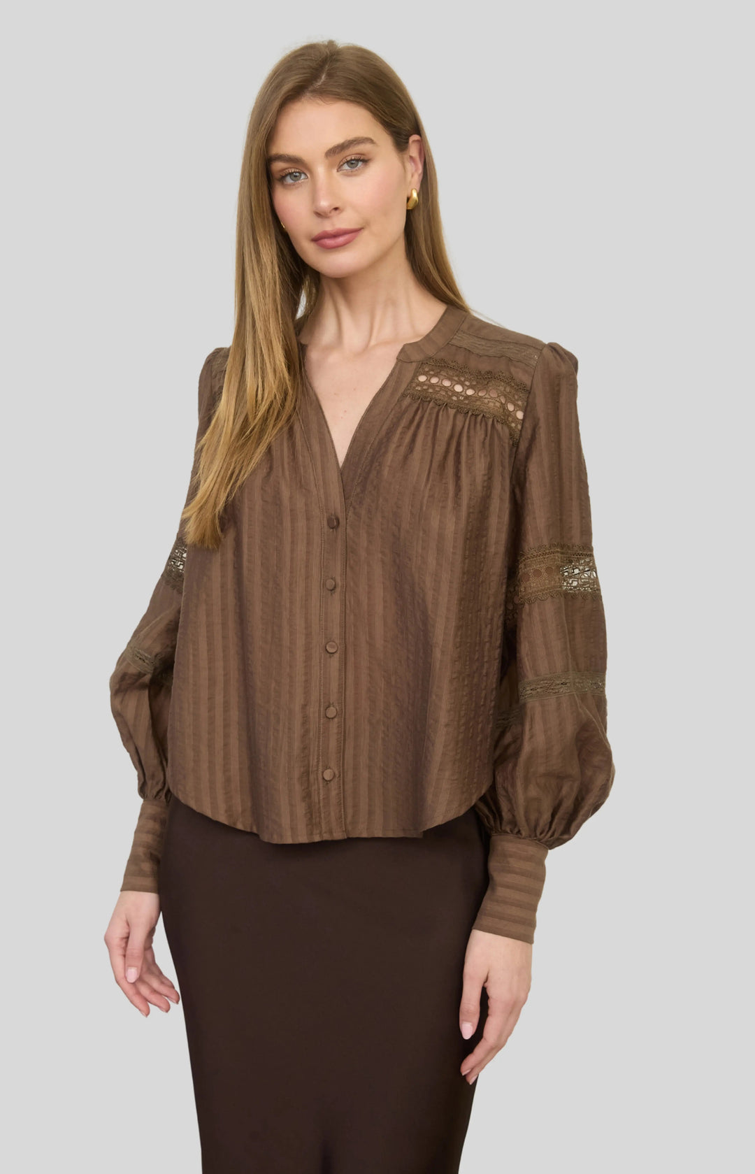 A woman modeling a brown long-sleeve button-front blouse with vertical textured stripes, gathered detailing at the neckline, and lace-like embroidery on the upper chest and sleeves, paired with dark brown pants against a light gray studio background.