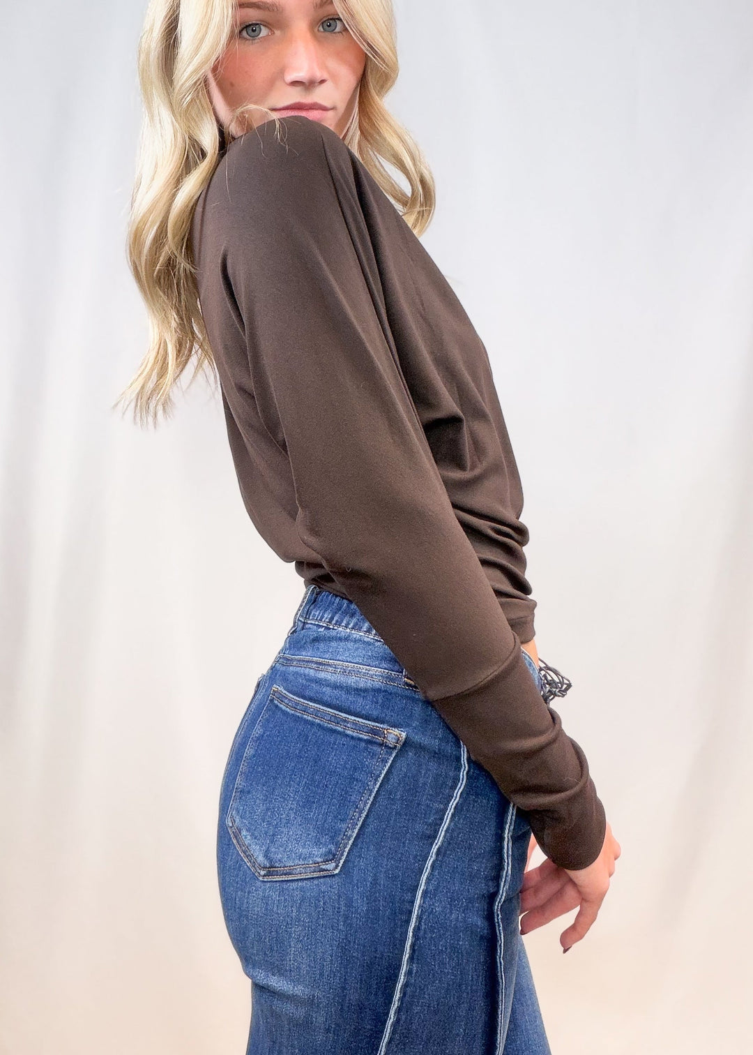 Woman wearing brown long sleeve top with denim jeans shown from side view on plain background