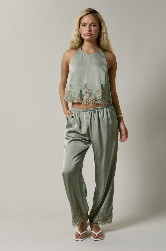 A woman modeling a sage green two-piece set consisting of a sleeveless crop top with floral lace trim and matching wide-leg pants with lace detailing at the hems, set against a plain light gray studio background.