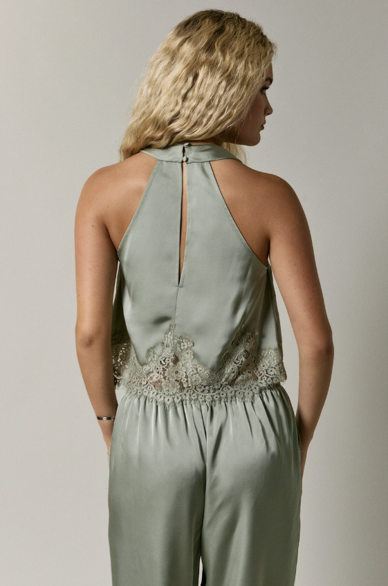 A woman shown from the back wearing a sage green halter jumpsuit with lace detailing at the waist, against a plain light gray studio background.