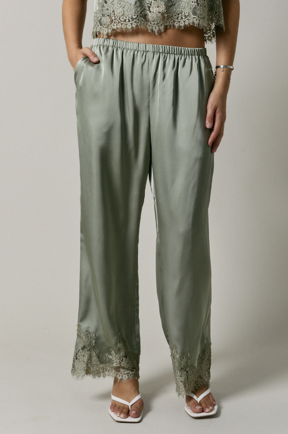 A person wearing wide-leg sage green satin pants with lace trim at the hem, paired with white heeled sandals, shown against a plain light gray studio background.